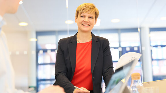 Vor einem Aktenschrank ist eine dunkelblonde Frau in dunkler Jacke und roter Bluse zu sehen.