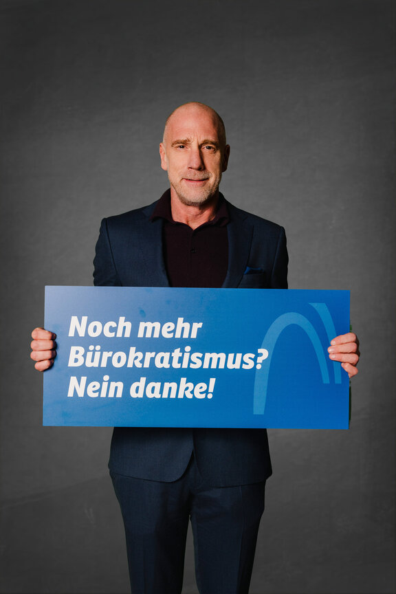 Eine Person steht vor einem neutralen grauen Hintergrund und hält ein großes blaues Schild mit der weißen Aufschrift: "Die Strafabgabe schwächt den Wirtschaftsstandort!"