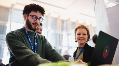 Zu sehen ist ein junger Mann mit kurzen, dunklen, lockigen Haaren. Er Trägt eine Brille und einen dunkelgrünen Pullover. Er steht an einem Tisch, darauf ein Laptop an dem er arbeitet. Neben ihm eine junge Frau, die beobachtet, was er am Laptop tut.