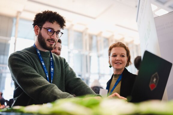 Zu sehen ist ein junger Mann mit kurzen, dunklen, lockigen Haaren. Er Trägt eine Brille und einen dunkelgrünen Pullover. Er steht an einem Tisch, darauf ein Laptop an dem er arbeitet. Neben ihm eine junge Frau, die beobachtet, was er am Laptop tut.