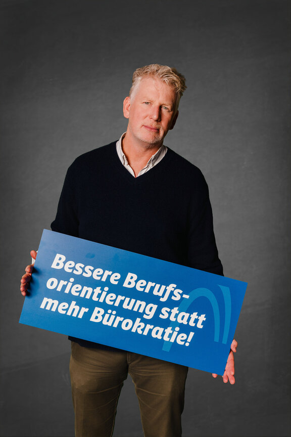 Eine Person steht vor einem neutralen grauen Hintergrund und hält ein großes blaues Schild mit der weißen Aufschrift: "Bessere Berufsorientierung statt mehr Bürokratie"