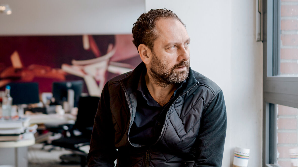 Ein Mann mittleren Alters mit Bart und dunkler Kleidung schaut aus einem Fenster.