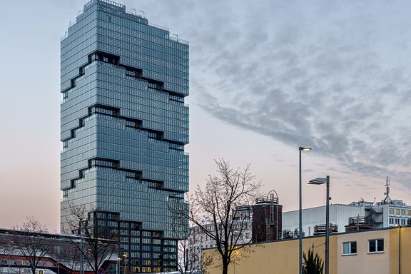 Hoher moderner Bürogebäudekomplex mit gestuften Glasfassaden bei Dämmerung, im Vordergrund Bäume und niedrige Gebäude