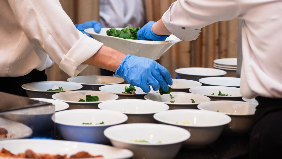 Zu sehen ist ein Tisch auf dem mehrere weiße Schüsseln stehen. Diese werden gerade mit Essen angerichtet.