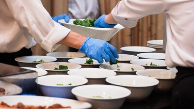 Zu sehen ist ein Tisch auf dem mehrere weiße Schüsseln stehen. Diese werden gerade mit Essen angerichtet.