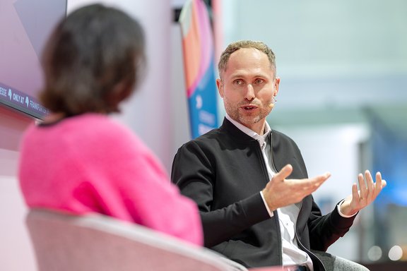 Zwei Personen sitzen sich gegenüber, eine trägt einen schwarzen Pullover und gestikuliert mit beiden Händen, die andere trägt ein pinkes Oberteil.