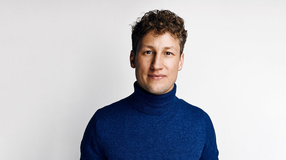 Mann mit lockigem Haar trägt einen blauen Rollkragenpullover vor hellem Hintergrund.