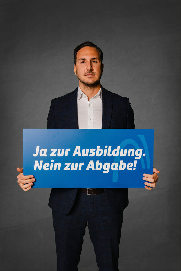 Eine Person steht vor einem neutralen grauen Hintergrund und hält ein großes blaues Schild mit der weißen Aufschrift: "Ja zur Ausbildung. Nein zur Abgabe!"