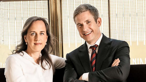 Zwei Personen sitzen nebeneinander, eine trägt einen schwarzen Anzug mit gestreifter Krawatte, die andere ein weißes Oberteil, im Hintergrund ein Fenster mit Jalousien.