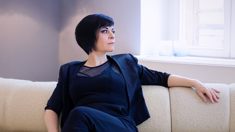 Zu sehen ist eine Frau mit mittellangen schwarzen Haaren auf einer hellen Couch sitzend.