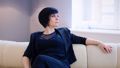Zu sehen ist eine Frau mit mittellangen schwarzen Haaren auf einer hellen Couch sitzend.