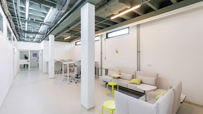 Heller, moderner Bürobereich mit weißen Wänden, zwei Sofas mit grünen Kissen und kleinen gelben Tischen, offenes Deckenraster mit sichtbaren Rohren