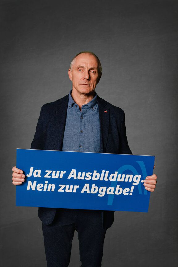 Eine Person steht vor einem neutralen grauen Hintergrund und hält ein großes blaues Schild mit der weißen Aufschrift: "Ja zur Ausbildung. Nein zur Abgabe!"