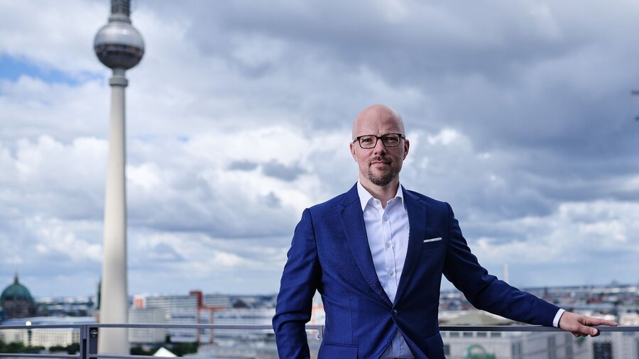 Zu sehen ist ein Mann im Anzug, welcher vor der Berliner Skyline steht und in die Kamera schaut.