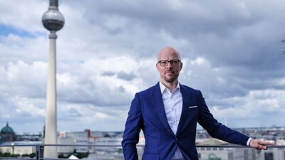 Zu sehen ist ein Mann im Anzug, welcher vor der Berliner Skyline steht und in die Kamera schaut.