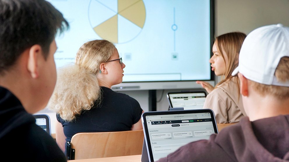 Vier Personen sitzen an Tischen mit Laptops, im Hintergrund ist eine Präsentation mit einem Kreisdiagramm auf einem Bildschirm zu sehen.