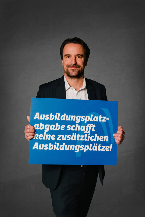 Eine Person steht vor einem neutralen grauen Hintergrund und hält ein großes blaues Schild mit der weißen Aufschrift: "Ausbildungsplatzabgabe schafft keine zusätzlichen Ausbildungsplätze!"