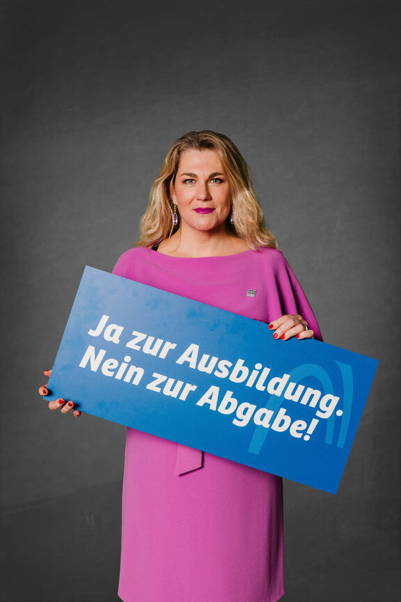 Eine Person steht vor einem neutralen grauen Hintergrund und hält ein großes blaues Schild mit der weißen Aufschrift: "Ja zur Ausbildung. Nein zur Abgabe!"
