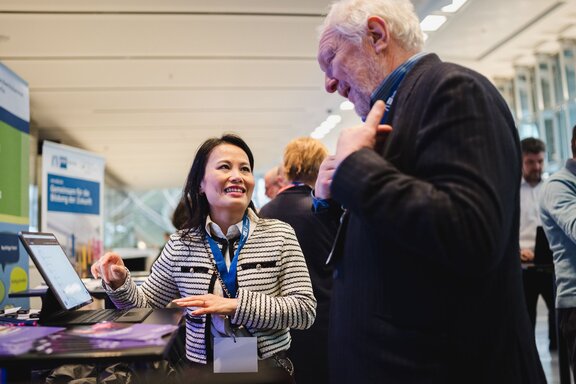 Zu sehen ist ein älterer Herr mit weißen kurzen Haaren, der ein dunkles Sakko trägt sowie eine mittelalte Frau mit halblangen schwarzen Haaren. Sie stehen an einem Tisch, auf dem ein kleiner Bildschirm zu sehen ist und unterhalten sich.