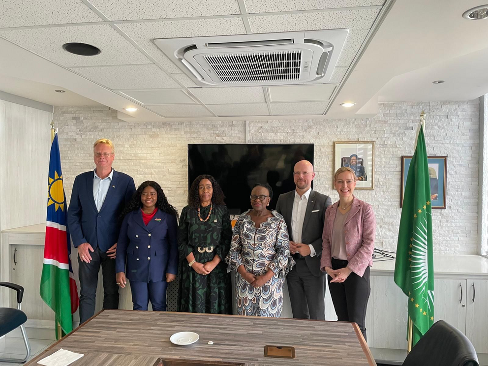 Eine Gruppe von sechs Personen steht in einem Konferenzraum nebeneinander. Hinter der Gruppe befinden sich ein großer schwarzer Bildschirm, eine weiße Wand und mehrere gerahmte Bilder. Links steht eine Flagge Namibias, rechts eine Flagge der Afrikanischen Union