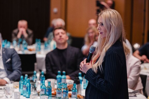 Zu sehen ist eine Blonde Frau mit einem Mikrofon in der Hand. Sie stellt eine Zwischenfrage während eine Rede gehalten wird.