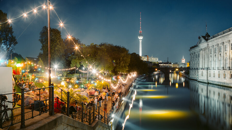 Belebte Außengastronomie am Berliner Spreeufer bei Nacht mit Lichterketten, vielen Gästen und Blick auf den Fernsehturm.