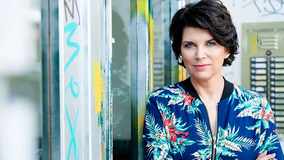Person mit verschränkten Armen trägt eine blaue Jacke mit floralem Muster und steht vor einer Metalltür mit Graffiti.