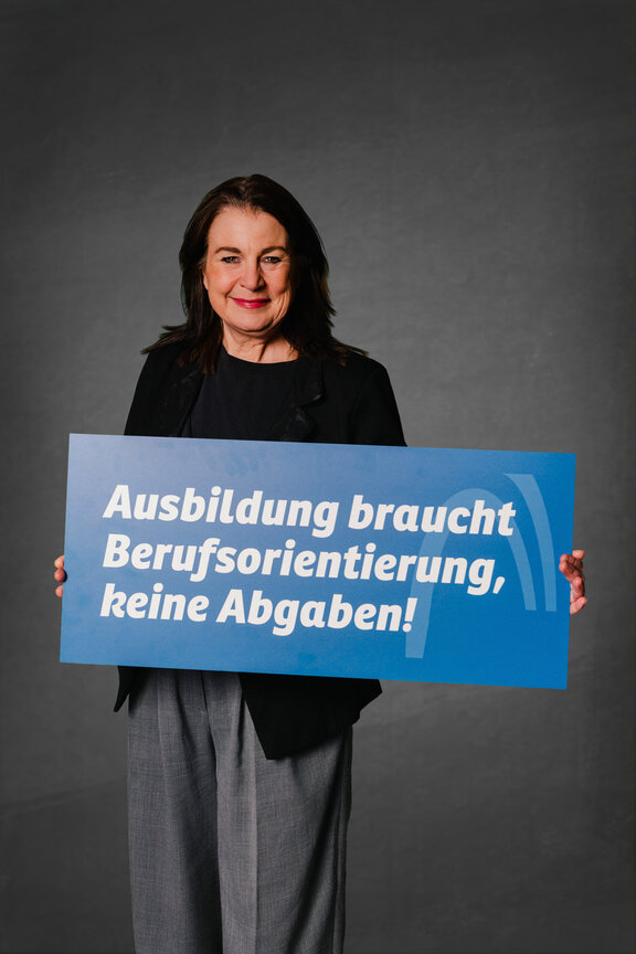 Eine Person steht vor einem neutralen grauen Hintergrund und hält ein großes blaues Schild mit der weißen Aufschrift: "Ausbildung braucht Berufsorientierung, keine Abgabe"