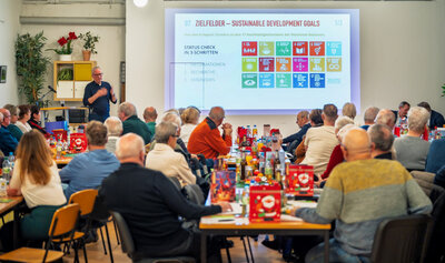 Auf dem Bild ist zusehen wie ein Mann mittleren Alters eine Präsentation hält. In dem Raum sitzen mehrere Menschen und  es sind nur Hinterköpfe zusehen.